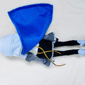 May include: A plush doll with a blue cape, white dress, black leggings, and blue and yellow shoes.
