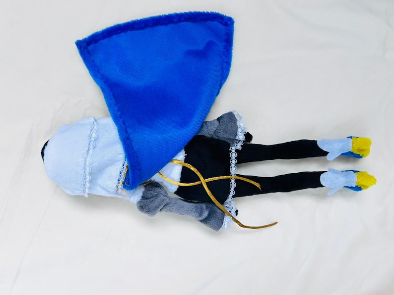 May include: A plush doll with a blue cape, white dress, black leggings, and blue and yellow shoes.