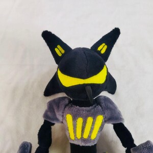 May include: A plush toy character with a black head and body, yellow detailing, and two pointed ears. The character has a yellow face and chest markings. The toy is made of soft, plush material.