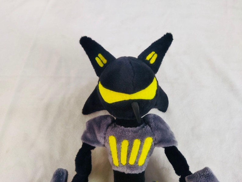 May include: A plush toy character with a black head and body, yellow detailing, and two pointed ears. The character has a yellow face and chest markings. The toy is made of soft, plush material.