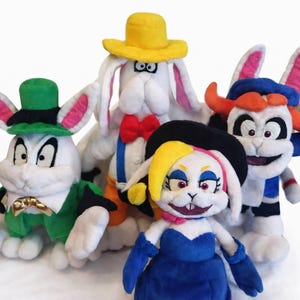 Peut inclure: Cinq poupées en peluche représentant des lapins, vêtues de tenues variées. L'un porte un chapeau et une veste verts, un autre un chapeau jaune et un nœud papillon, et un troisième une robe bleue et un chapeau noir. Les lapins ont une fourrure blanche et des oreilles roses.