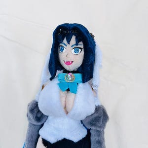 May include: A plush toy of a female anime character with long black hair, wearing a blue and white dress with a blue bow and a jeweled collar. The toy has a white background.