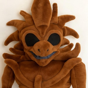 May include: A brown plush creature with large black eyes, a smiling mouth, and spiky details. The soft toy has a unique design, with a long, slender appendage extending from its chest. The plushie is made of a soft, fuzzy material.