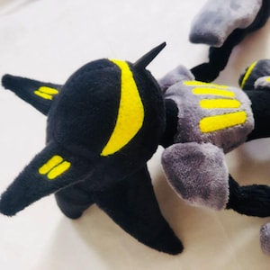 May include: A black, grey, and yellow plush robot toy. The toy features a black head with yellow markings and a segmented body. The body is grey with yellow detailing.