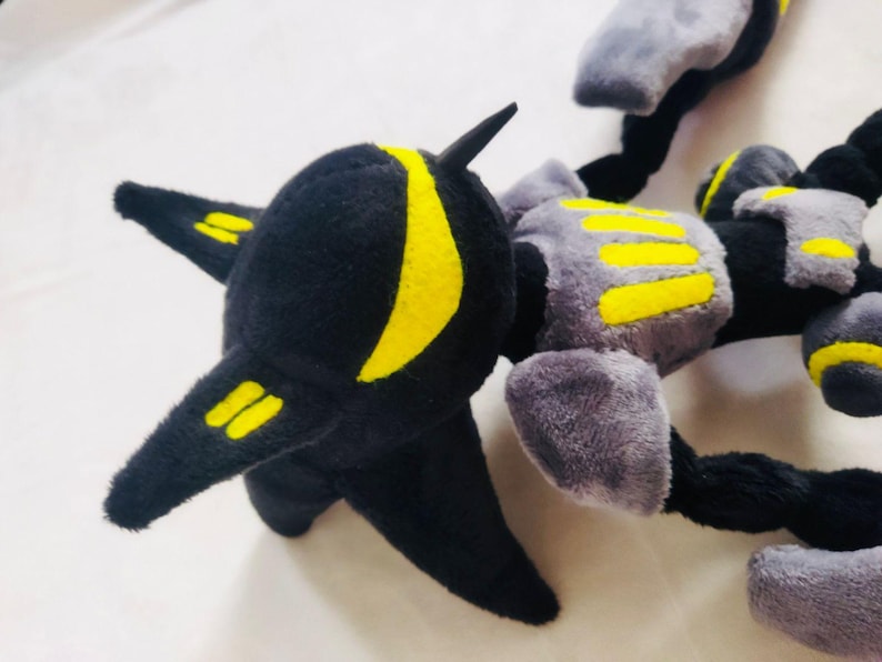 May include: A black, grey, and yellow plush robot toy. The toy features a black head with yellow markings and a segmented body. The body is grey with yellow detailing.
