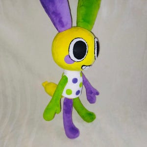 Puede incluir: Un peluche de conejito con cabeza amarilla, orejas moradas y verdes, y una camisa blanca con lunares verdes y morados. El conejito tiene ojos negros y una pequeña cola amarilla. Los brazos y las piernas son verdes y moradas.
