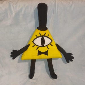 Könnte beinhalten: Ein gelbes, dreieckiges Plüschtier mit einem einzelnen Auge, schwarzen Armen, Beinen und einem Zylinder. Das Auge ist weiß mit einer schwarzen Pupille. Eine schwarze Fliege befindet sich unter dem Auge. Das Spielzeug ist eine Figur aus der Zeichentrickserie Gravity Falls.