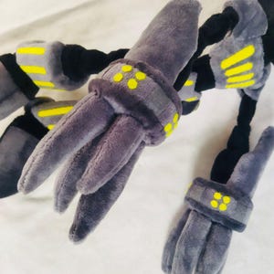 May include: A plush toy with a robotic design, in shades of grey, black, and yellow. The toy has multiple articulated limbs with grey, finger-like appendages. Yellow details are present on the joints and bands.