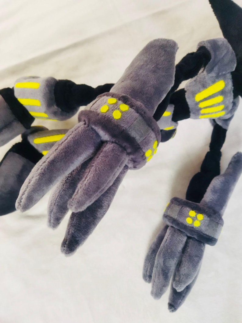 May include: A plush toy with a robotic design, in shades of grey, black, and yellow. The toy has multiple articulated limbs with grey, finger-like appendages. Yellow details are present on the joints and bands.
