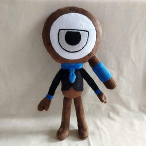 May include: A plush toy in the shape of a cartoon character with a large eye. The toy has a brown body, a white and black eye, and wears a black jacket, blue scarf, and blue armbands. The toy is approximately 12 inches tall.