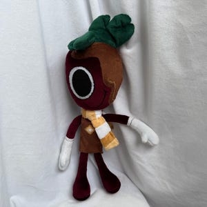 Pode incluir: Boneca de pelúcia em forma de personagem semelhante a um rabanete. A boneca tem cabeça e pernas bordô, cabelo castanho e uma parte superior verde folhosa. Usa um colete marrom e um cachecol amarelo e branco. Tem mãos brancas e um grande olho preto.