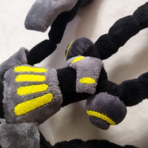 May include: Close-up of a plush toy with black, grey, and yellow accents. The toy has segmented black limbs with grey padded sections and yellow stripes. The plush appears soft and textured.