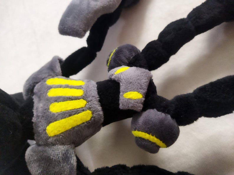 May include: Close-up of a plush toy with black, grey, and yellow accents. The toy has segmented black limbs with grey padded sections and yellow stripes. The plush appears soft and textured.