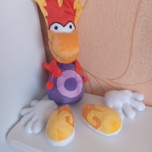 Puede incluir: Un juguete de peluche de un pájaro rojo y amarillo con un cuerpo morado y manos blancas. El pájaro tiene un pico naranja grande y una cresta amarilla.