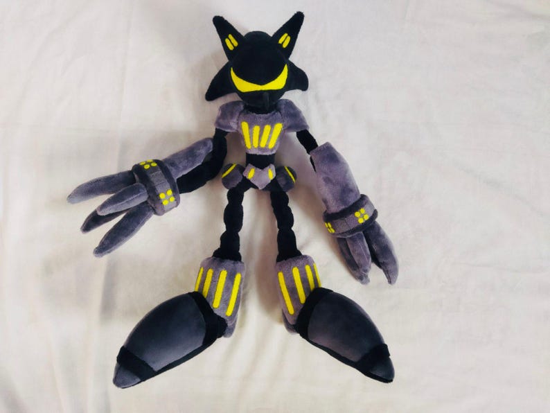 May include: A plush toy robot character in black, grey, and yellow. The toy has a distinctive head shape, large hands, and feet. The robot's body features yellow accents and is posed on a white surface.