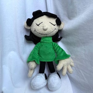 May include: A soft toy doll depicting a cartoon character. The doll has black hair and a black hat. It wears a green turtleneck jumper, black trousers, and white shoes. The face features closed eyes and a stitched smile.