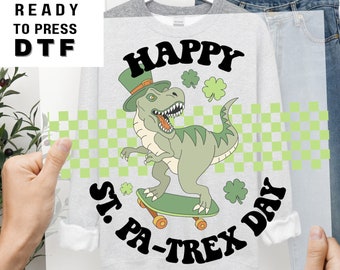 Puede incluir: Una sudadera gris con un dinosaurio verde que lleva un sombrero verde y monta en patinete. El dinosaurio está rodeado de tréboles verdes y el texto "Happy St. Pa-trex Day".