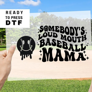 May include: A black and white design on a clear sheet, featuring a smiley face with baseball stitches for eyes, dripping down, and the text "Somebody's Loud Mouth Baseball Mama" in a retro font.