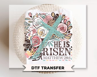 May include: White t-shirt featuring a floral design with pink, yellow, and brown accents, and the text "HE IS RISEN" with "MATTHEW 28:6". A teal cross is superimposed over the design. The words "DTF TRANSFER" are at the bottom.