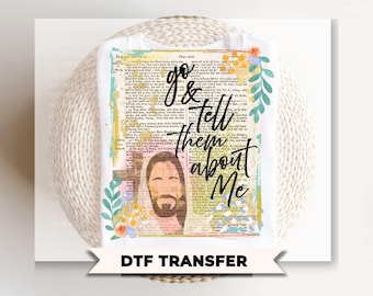 May include: White t-shirt featuring a design with the text "go & tell them about Me" in black script. The design includes an image of Jesus and is set against a vintage book page background with floral accents. The words "DTF TRANSFER" are at the bottom.