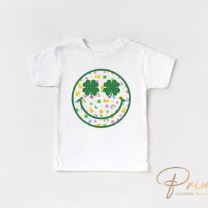 May include: White toddler t-shirt featuring a green smiley face design with shamrock eyes and a colorful pattern. The design includes rainbows, pots of gold, and other St. Patrick's Day themed graphics. The shirt is a short-sleeved crew neck.