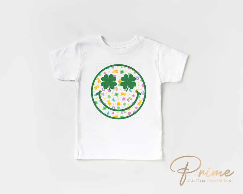 May include: White toddler t-shirt featuring a green smiley face design with shamrock eyes and a colorful pattern. The design includes rainbows, pots of gold, and other St. Patrick's Day themed graphics. The shirt is a short-sleeved crew neck.