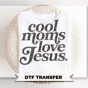 May include: White t-shirt with the text "cool moms love Jesus" in a dark gray, stylized font. Below the shirt is a banner that reads "DTF TRANSFER". The shirt is displayed on a woven, light-colored surface.