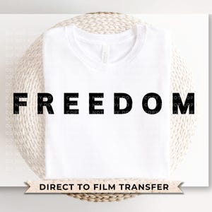 May include: A white t-shirt with the word "FREEDOM" printed in large, black letters. Below the shirt is a banner that reads "DIRECT TO FILM TRANSFER". The shirt is displayed on a white surface.