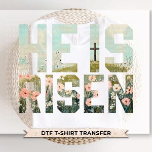 May include: White t-shirt with the words "HE IS RISEN" in large, stylized letters. The letters are filled with a watercolor design of a cross on a hill and floral elements. The words "DTF T-SHIRT TRANSFER" are at the bottom.