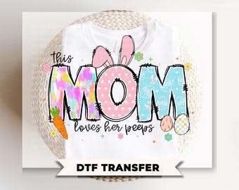 May include: White t-shirt with the words "this MOM loves her peeps" in colorful letters. The "MOM" is decorated with a pink polka dot "O" and the "M" is tie-dye. Easter eggs and a carrot are also featured.