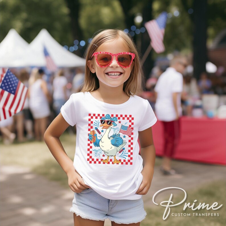 May include: A white t-shirt with a cartoon image of a goose wearing sunglasses and a red, white, and blue bandana. The goose is holding a sparkler and a small American flag. The image is surrounded by a red and white checkered border.