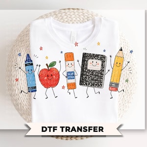 May include: A white t-shirt featuring a colorful graphic of anthropomorphic school supplies including a blue crayon, a red apple, an orange glue stick, a black and white composition notebook, and a yellow pencil, all surrounded by small stars.