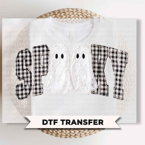 May include: A white t-shirt featuring a graphic design with the word SPOOKY in black and white buffalo plaid letters. Two white lace-textured ghost figures replace the letter O. A banner at the bottom displays the text DTF TRANSFER in black.