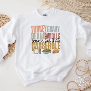 May include: White sweatshirt with a Thanksgiving themed graphic that says "Turkey Gravy Beans and Rolls lemme see that Casserole". The graphic features a turkey, a casserole dish, and a roll.