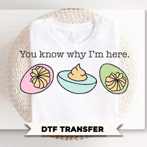 May include: White t-shirt with the text "You know why I'm here." and a graphic of three deviled eggs. The eggs are pink, blue, and green, with yellow filling. The bottom of the image reads "DTF TRANSFER".