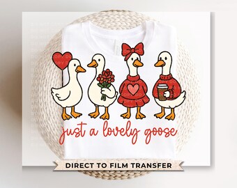 Peut inclure: T-shirt blanc avec un dessin animé de quatre oies, chacune avec des accessoires uniques : un ballon cœur, des fleurs, un nœud et une tasse de café. Le texte "just a lovely goose" est écrit en script rouge. Les mots "DIRECT TO FILM TRANSFER" sont également présents.