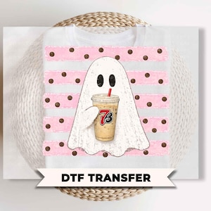 May include: A white t-shirt featuring a graphic of a ghost holding a coffee cup with a red straw. The ghost is centered over horizontal pink stripes and brown polka dots. The coffee cup displays the red and black text 73. The design is a DTF transfer.