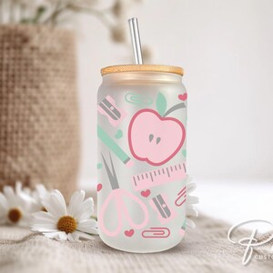 May include: A clear glass tumbler with a pink apple, scissors, pencil, ruler, and paper clips design. The tumbler has a wooden lid and a straw.