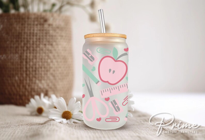 May include: A clear glass tumbler with a pink apple, scissors, pencil, ruler, and paper clips design. The tumbler has a wooden lid and a straw.
