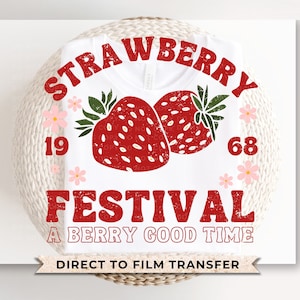 May include: White t-shirt with a red graphic that reads "STRAWBERRY FESTIVAL" above two red strawberries and the text "A BERRY GOOD TIME". The shirt also has the numbers "1968" and pink flower accents.