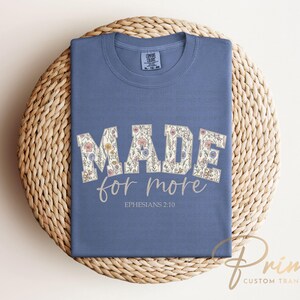 Puede incluir: Camiseta azul con la frase "MADE for more" en letras florales. El texto "Ephesians 2:10" est&aacute; impreso debajo. La camiseta est&aacute; doblada sobre una estera redonda tejida.