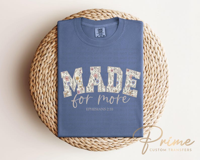 Puede incluir: Camiseta azul con la frase "MADE for more" en letras florales. El texto "Ephesians 2:10" est&aacute; impreso debajo. La camiseta est&aacute; doblada sobre una estera redonda tejida.