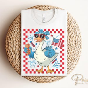 May include: A white t-shirt with a red, white, and blue checkered background. A cartoon goose wearing sunglasses and a red, white, and blue bandana holds a hot dog and a small American flag. The goose is surrounded by fireworks.