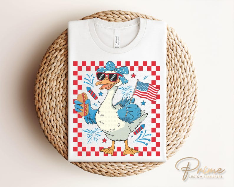 May include: A white t-shirt with a red, white, and blue checkered background. A cartoon goose wearing sunglasses and a red, white, and blue bandana holds a hot dog and a small American flag. The goose is surrounded by fireworks.