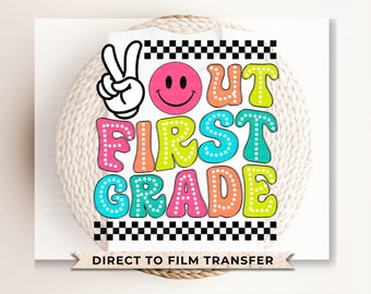 May include: A white t-shirt featuring a graphic design with a black and white checkered border, a peace sign hand gesture, a pink smiley face, and the colorful text "OUT FIRST GRADE" in a bubbly font with white polka dot accents throughout.