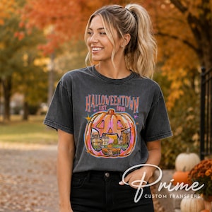 May include: A charcoal gray short-sleeve t-shirt featuring a graphic of a large orange jack-o'-lantern. Inside the pumpkin is a scene of a building and trees. The text reads HALLOWEENTOWN EST. 1998 with small colorful stars surrounding the design.