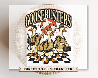 May include: White t-shirt featuring a humorous design with the text "GOOSERUSTERS" above a group of cartoon geese dressed as ghostbusters. The design includes a ghost and a "no ghost" logo. Below the design is the text "DIRECT TO FILM TRANSFER".