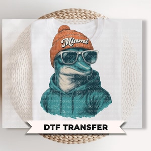 May include: A white DTF transfer featuring a graphic illustration of a dolphin wearing an orange knit beanie with the word Miami written in white script, dark sunglasses, and a teal hooded sweatshirt. The design has a distressed texture.