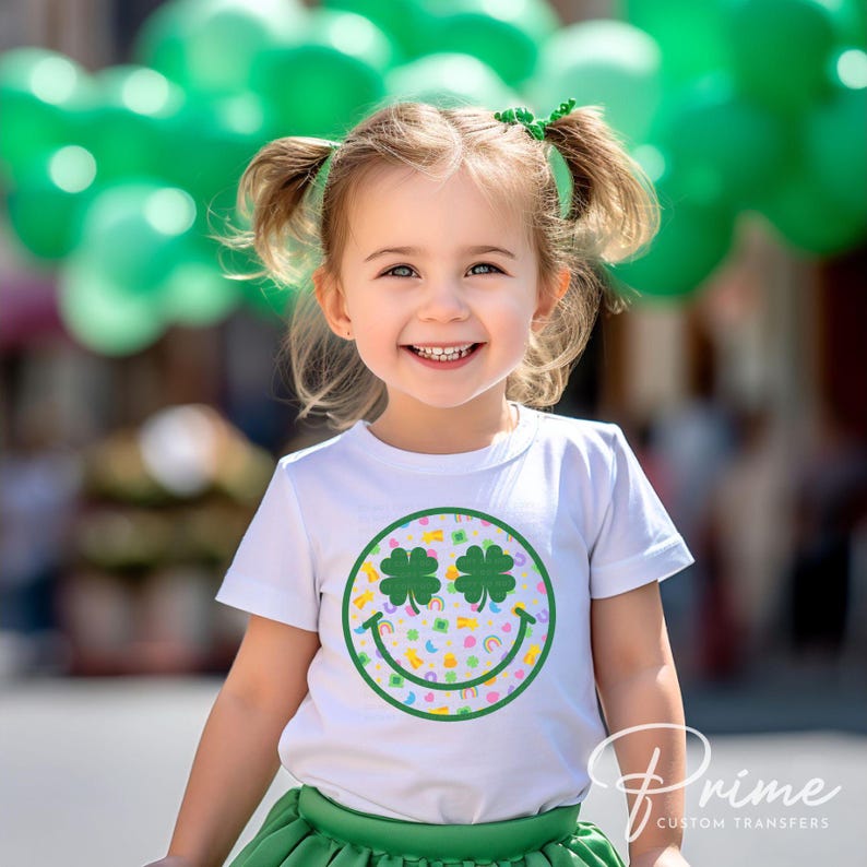 May include: A young child wearing a white t-shirt with a St. Patrick's Day design. The design features a green smiley face with shamrock eyes and colorful rainbows. The child is also wearing a green skirt and has green hair accessories.