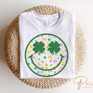 May include: White t-shirt featuring a St. Patrick's Day smiley face design. The smiley face has shamrock eyes and a pattern of rainbows, pots of gold, and shamrocks. The shirt is made of a soft material.